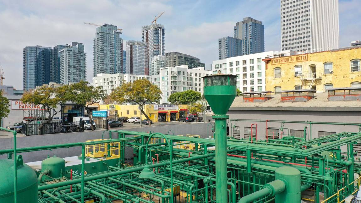 A petroleum well operation consisting of a network of green pipes, sits in the foreground of the Los Angleles downtown cityscape.
