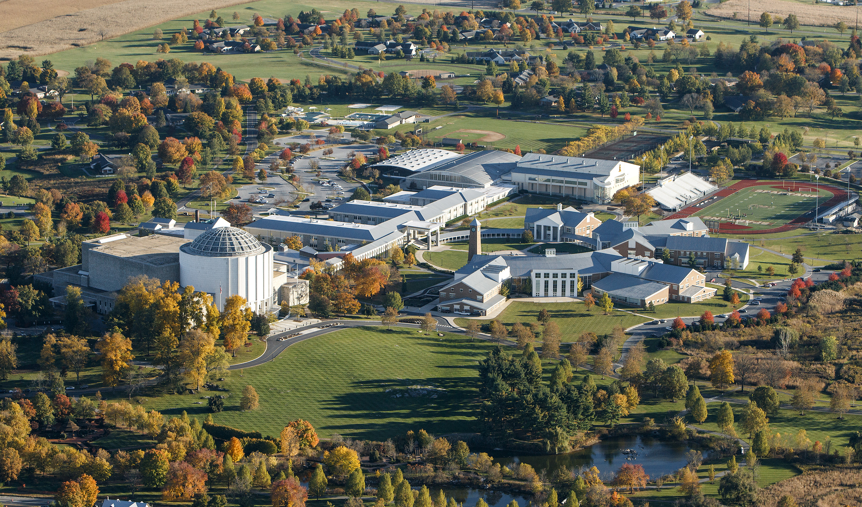 An aerial view of a campus of white and brick buildings spread out over rolling fields and surrounded by trees.