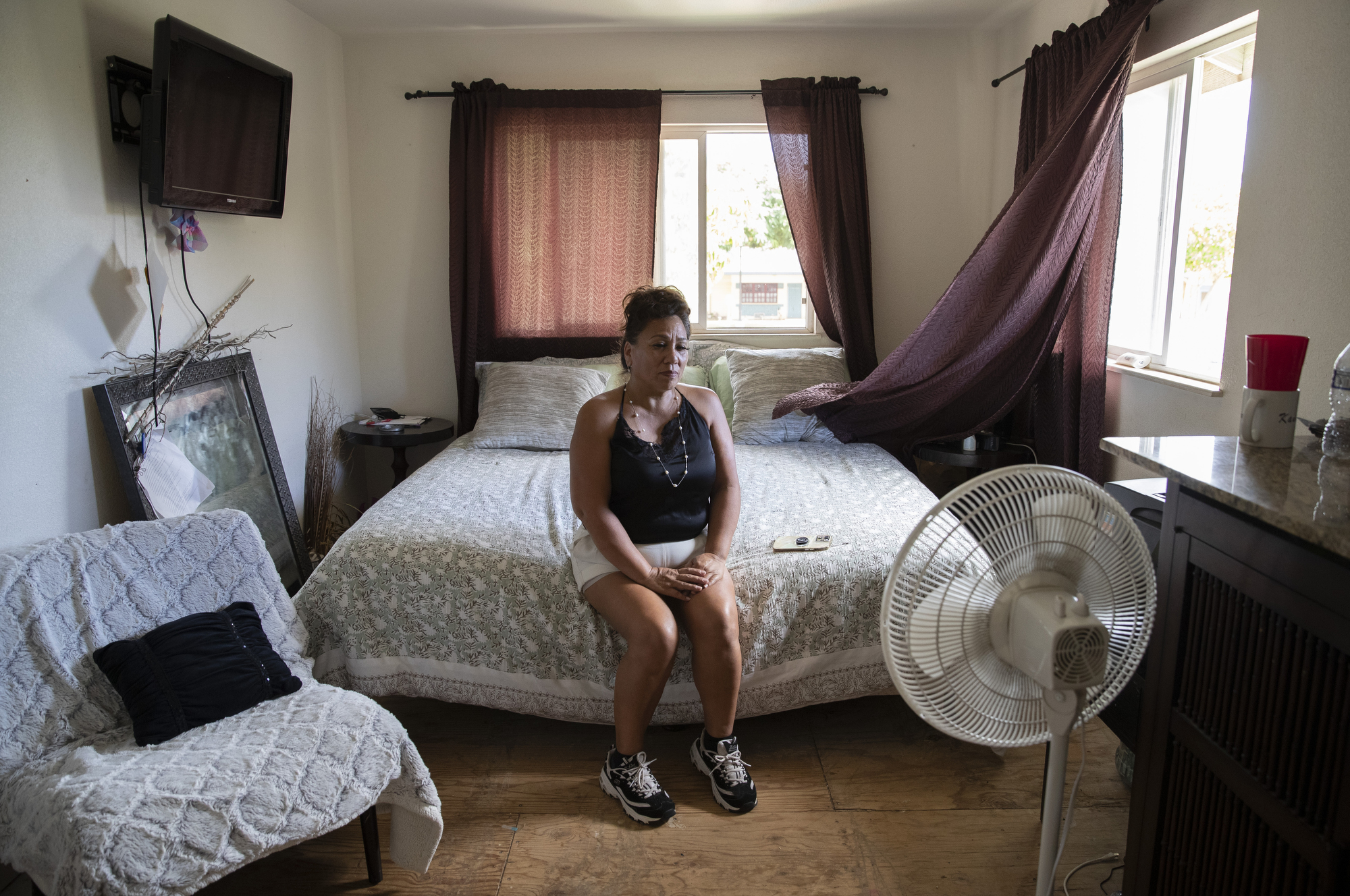 A woman sits on a bed in a room with a bare plywood floor.