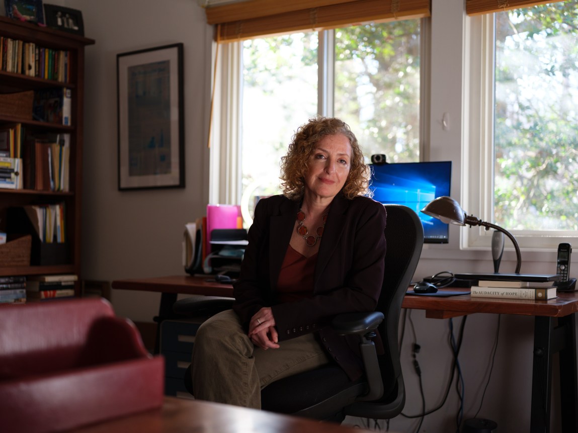 A white woman with curly blond hair sits in front of a desk by a window. She's wearing a blazer and a chunky orange necklace.