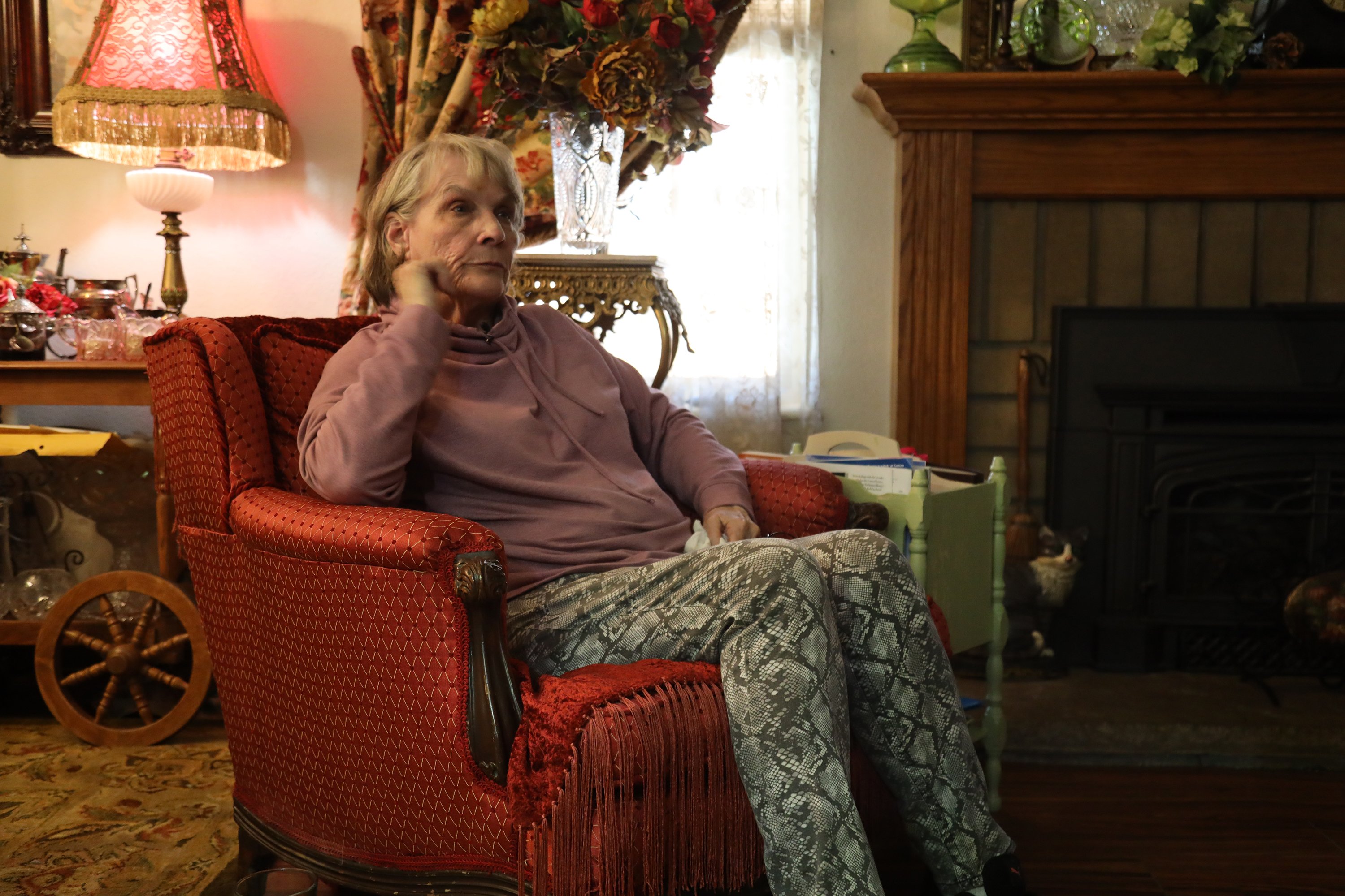 An older woman sits in an upholstered armchair in a room full of knickknacks.