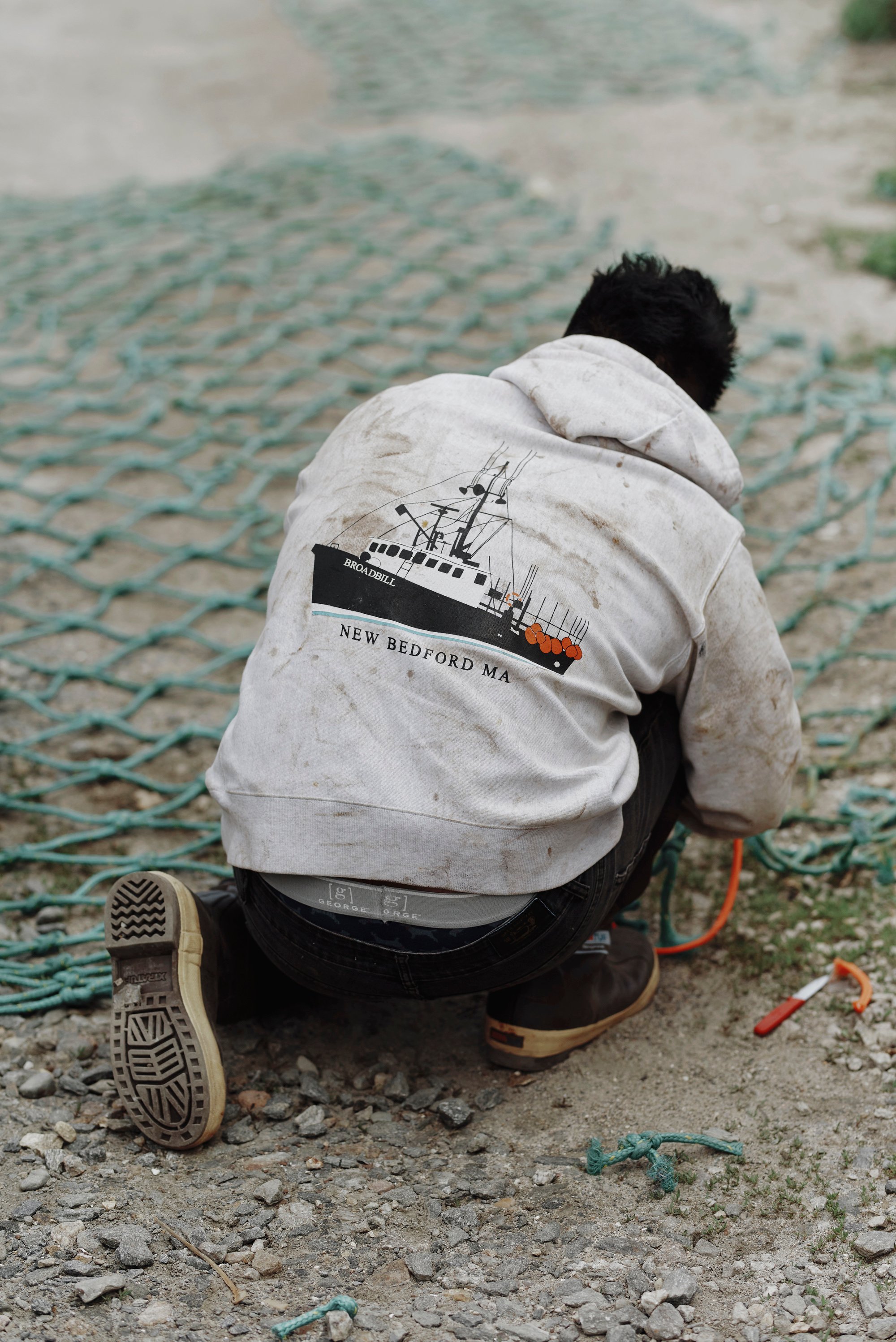 A man in a hoodie, seen from the back, crouches over a net on the beach.