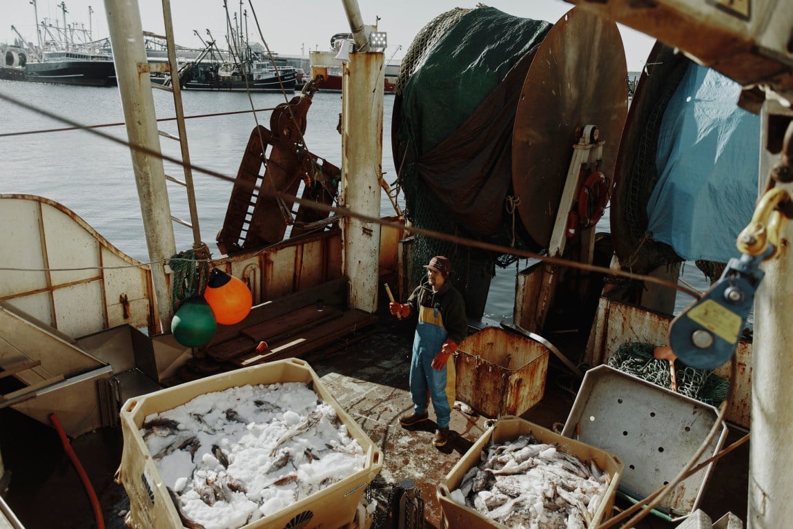 A man in waterproof overalls stands on the deck of a ship surrounded by crates, nets, buoys, tarps and other fishing apparatus.