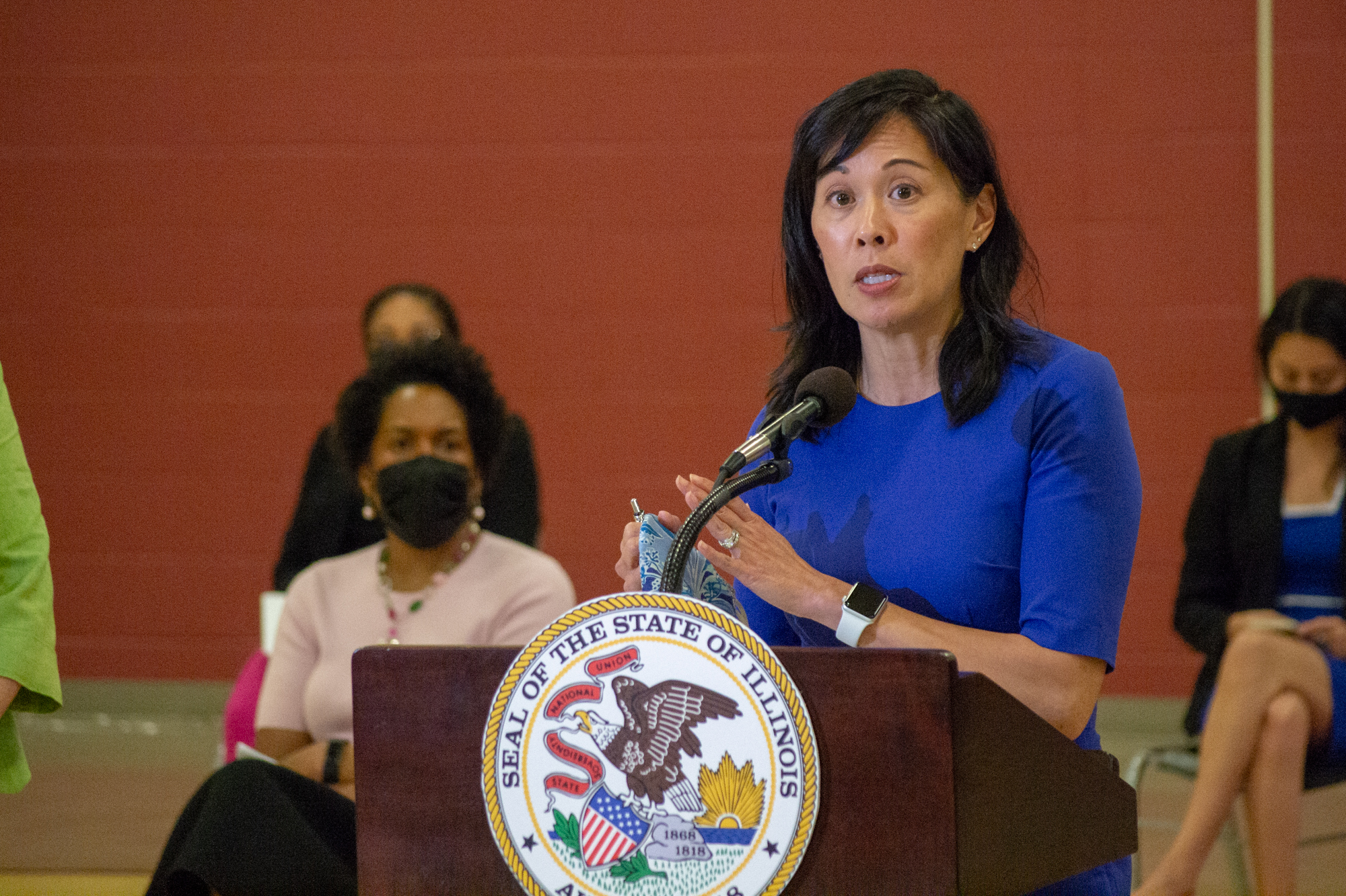 An Asian woman in a blue dress stands behind a lectern bearing the seal of the state of Illinois