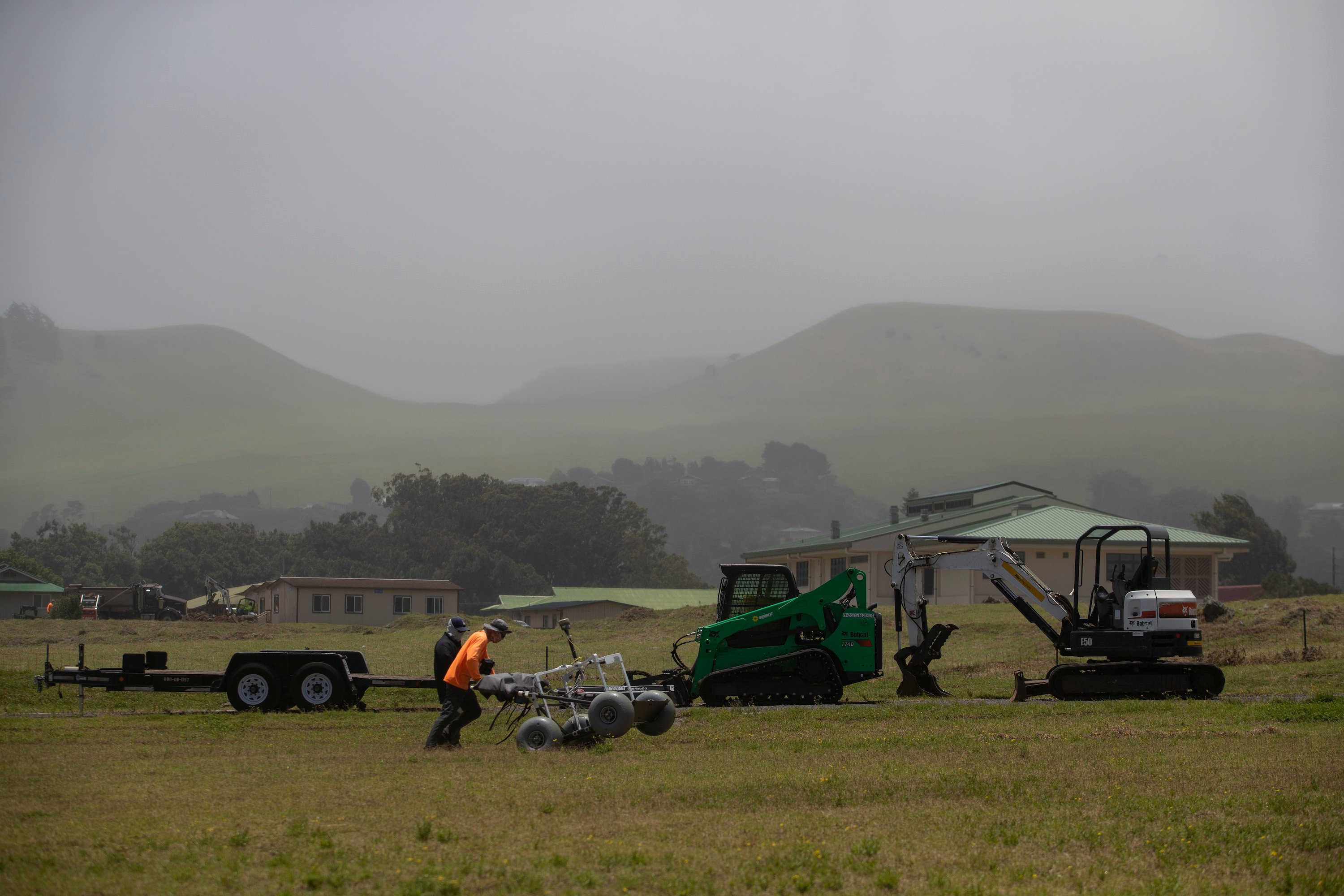 A man in an orange shirt and baseball cap pushes a four-wheeled device with balloon tires across a field.