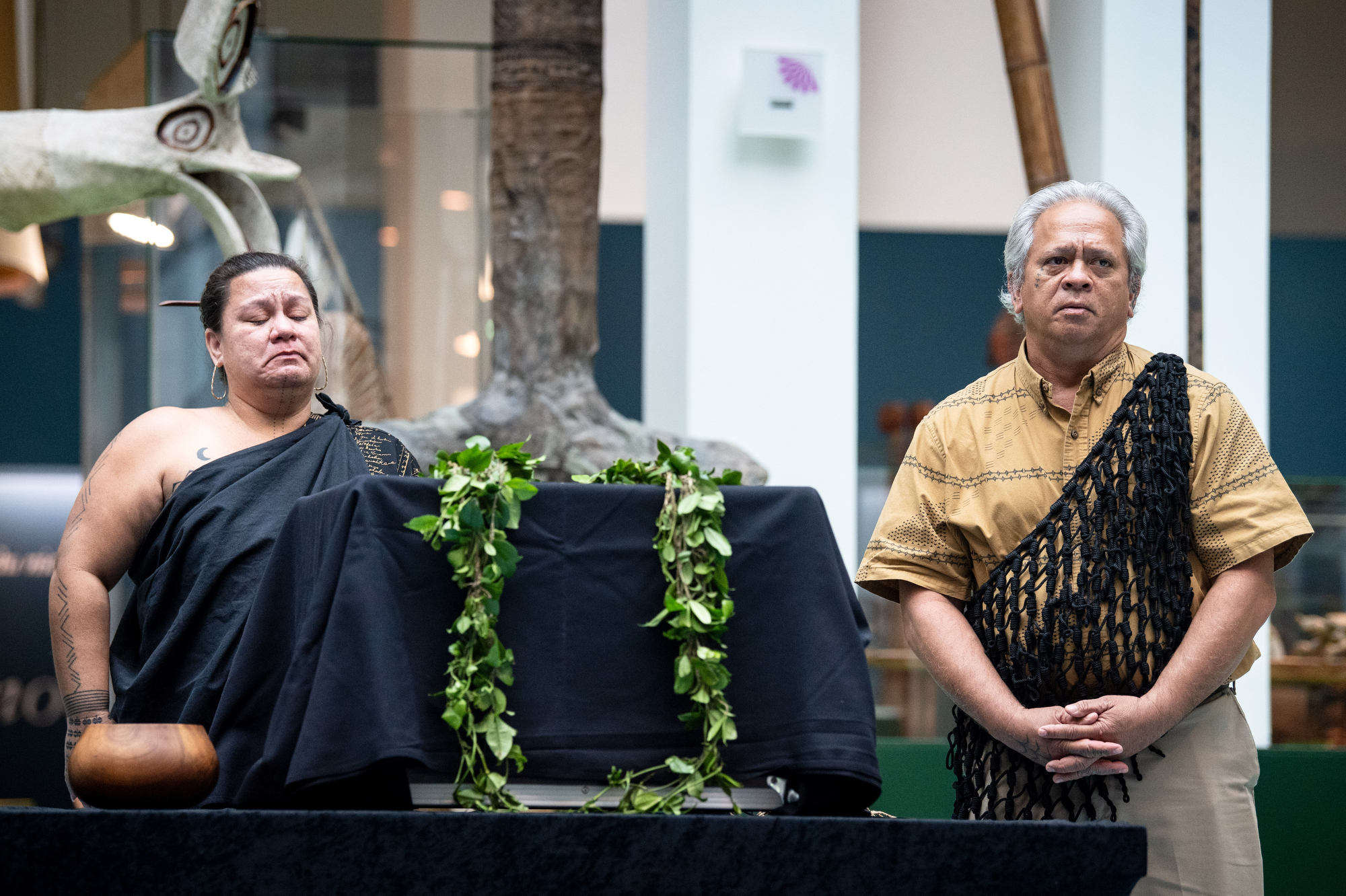 Two people stand on either side of a container covered in a cloth and wreathed with leaves.