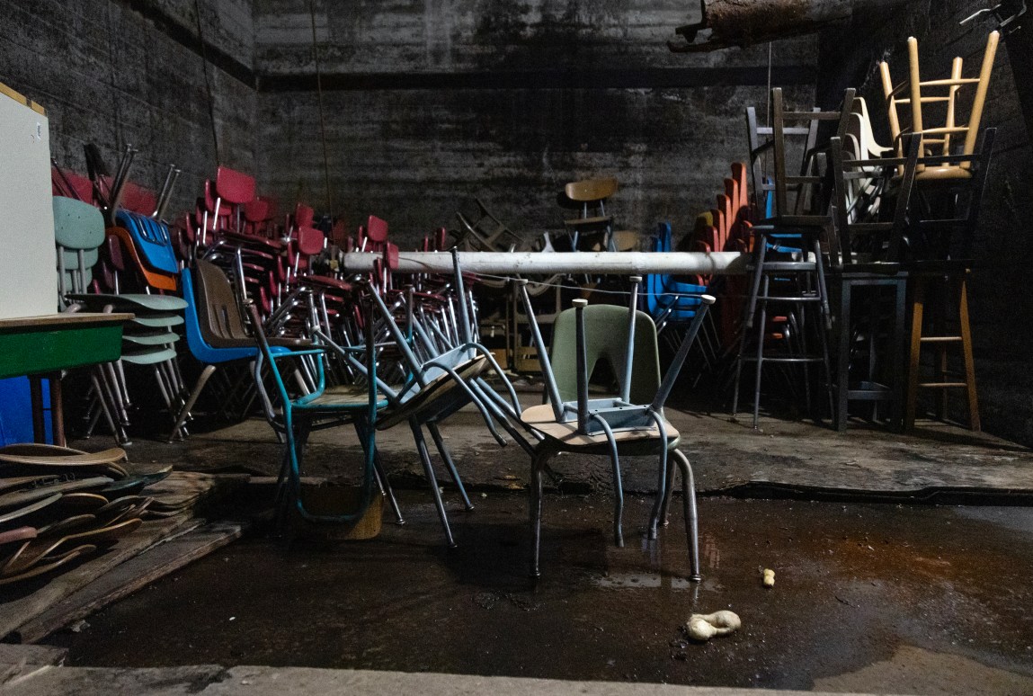 Many chairs stacked in a dark, unfinished space with puddles of water on the bare concrete floor.