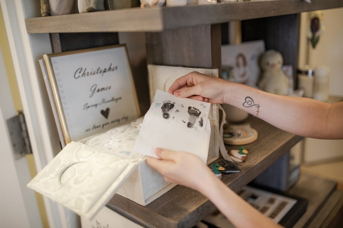 A white woman's hands hold a piece of paper with two small footprints inked on it. In the background, a framed paper says 