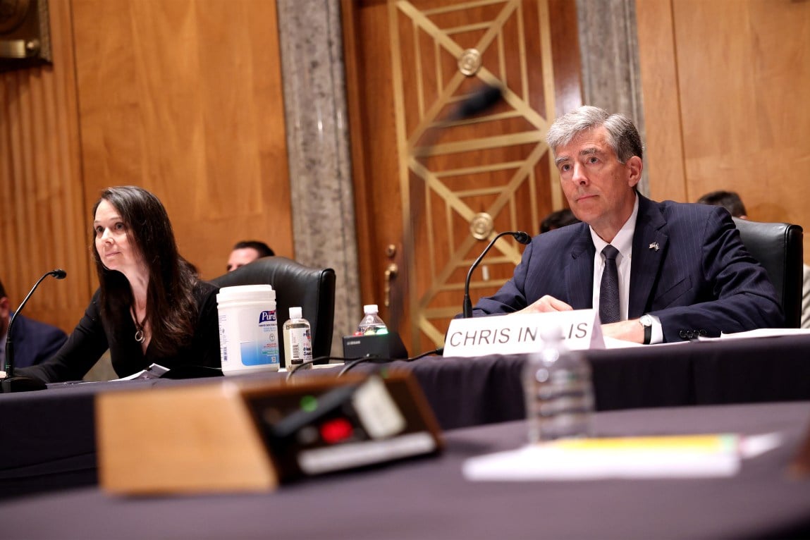 A man in a suit and tie with graying hair sits behind a nameplate reading 