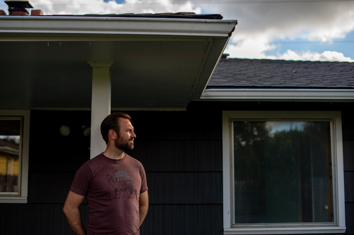 A bearded man in a brown t-shirt stands against a building and looks off to one side.