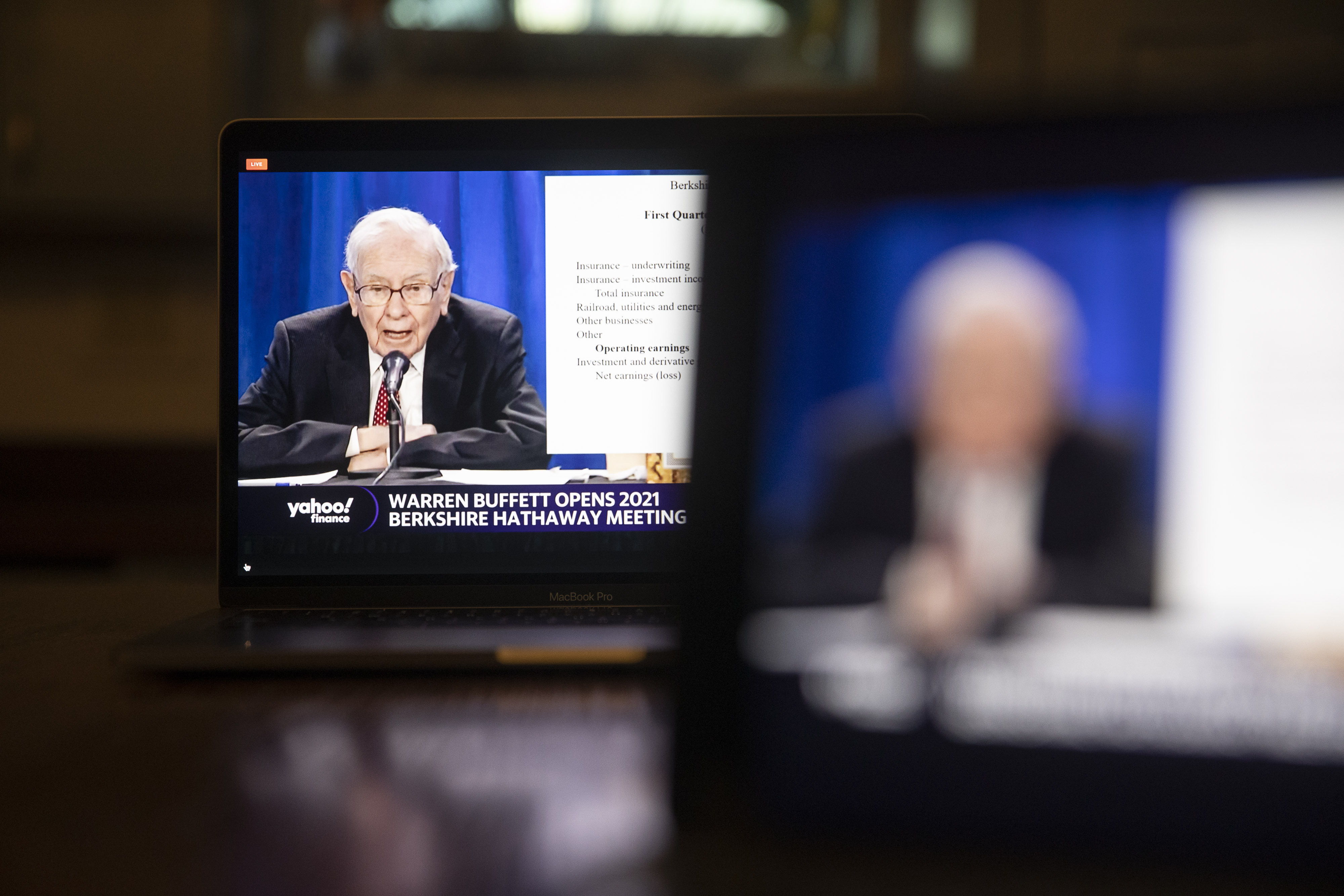 A man with white hair and glasses, wearing a suit, gestures while sitting at a table in front of a backdrop reading 