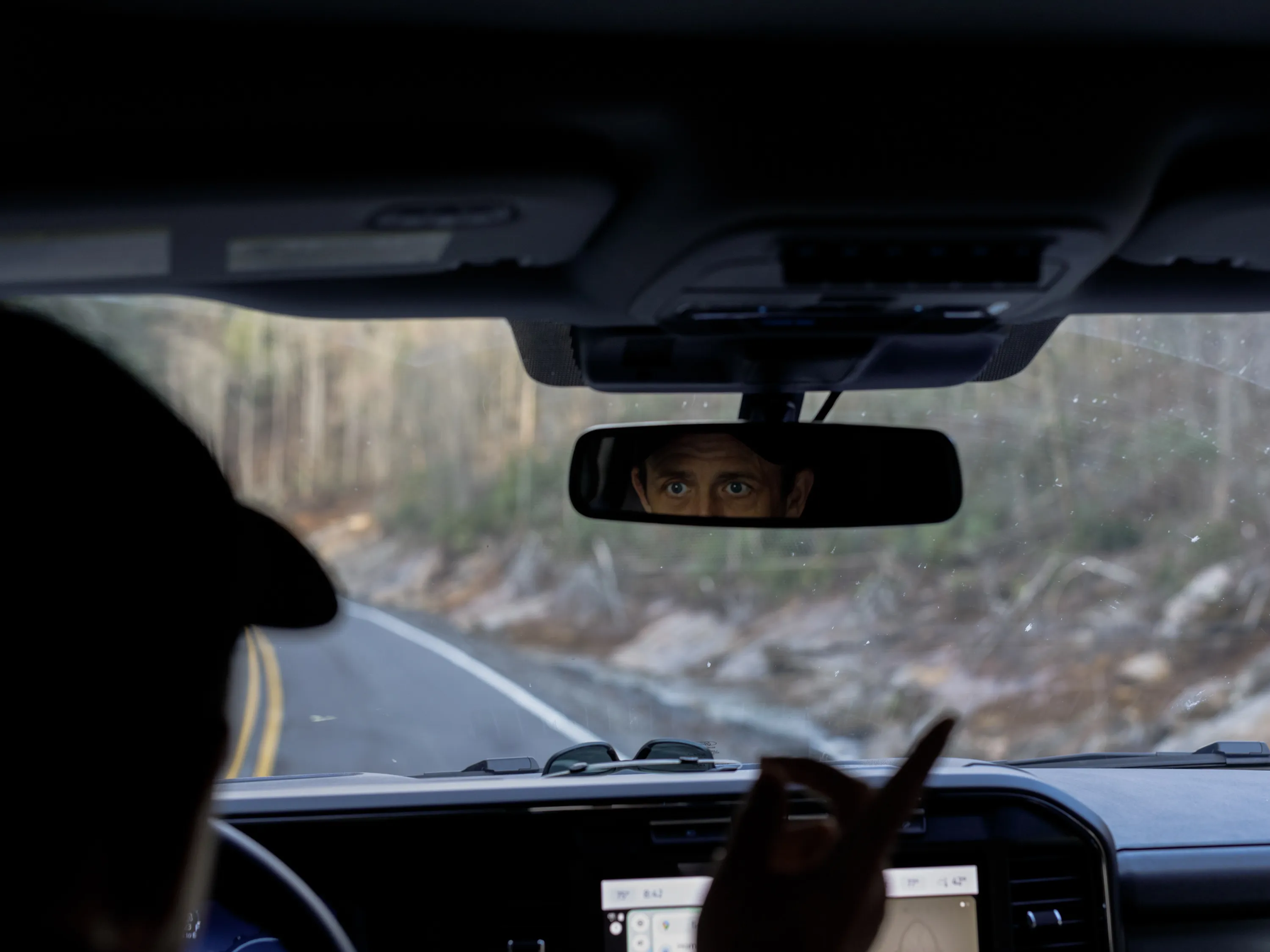 A male driver looks into a car’s rearview mirror as he points at the road outside the windshield.