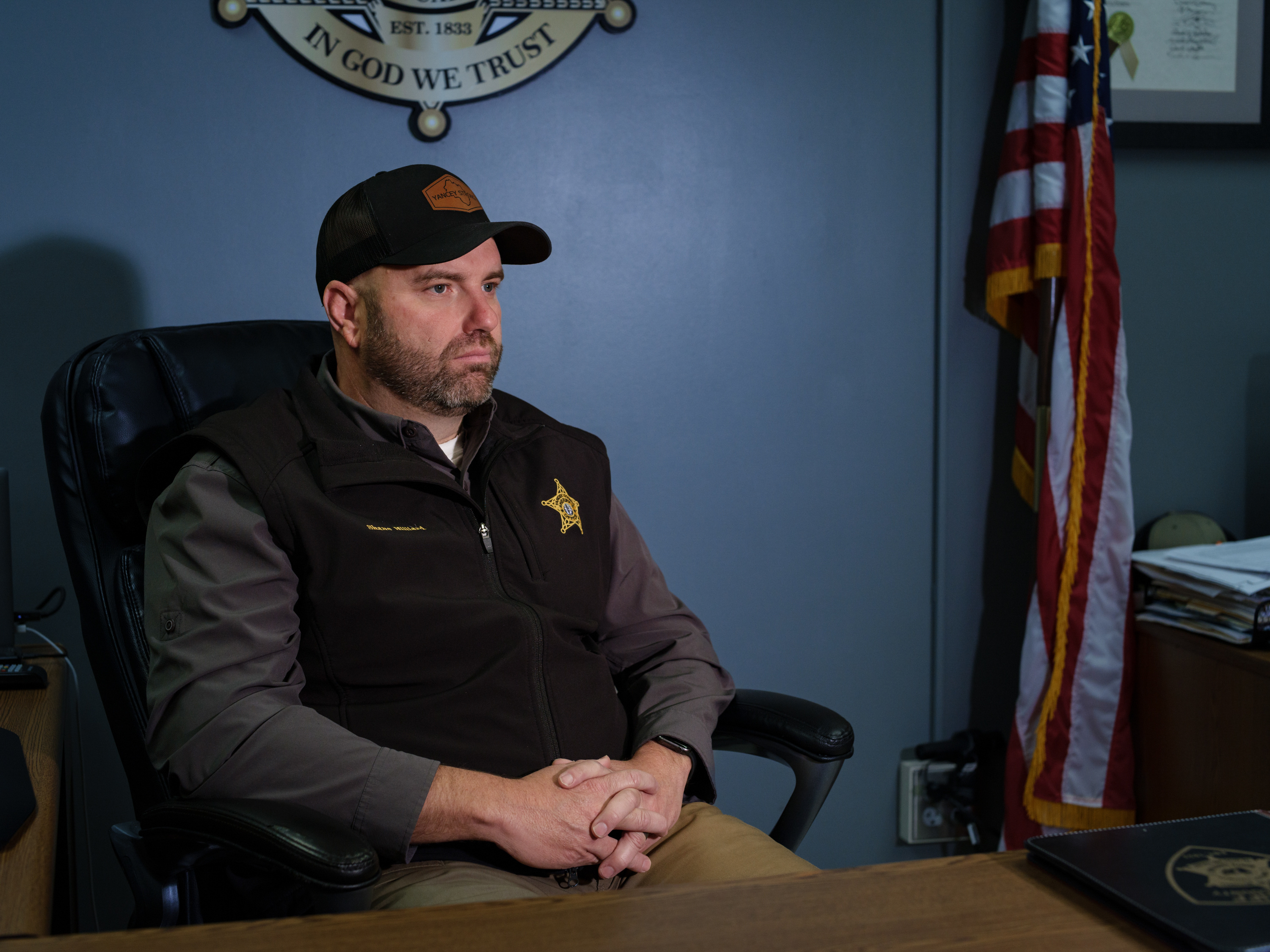 A man wearing a vest with a sheriff’s star sits in a desk chair next to an American flag and a sign with the text “In God We Trust.”