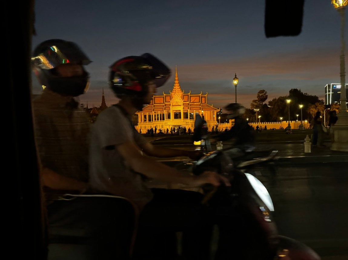 Two motorcyclists drive by a lit up Cambodian building.