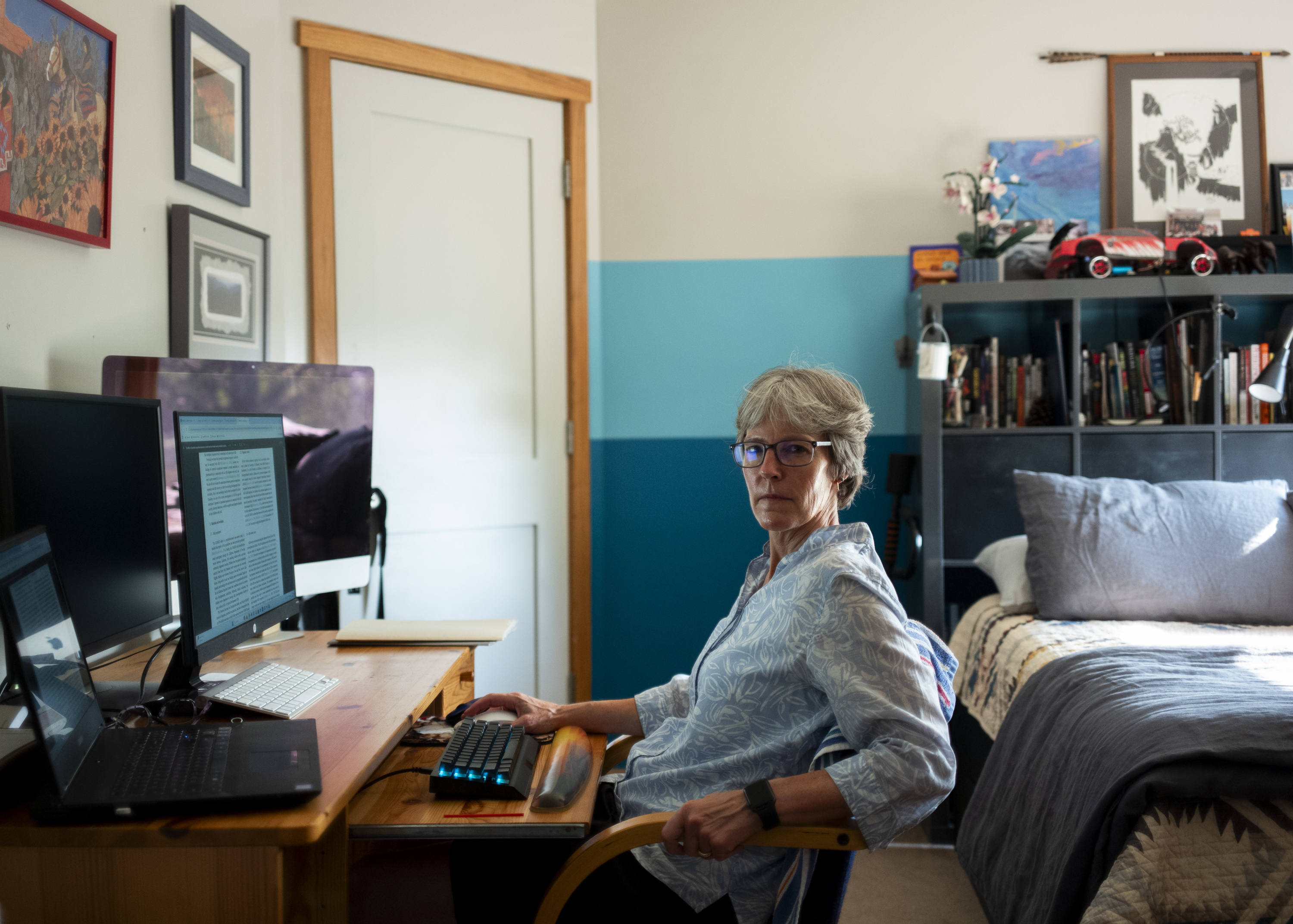 McCanlies sits at a wooden desk with multiple monitors and keyboards. Behind her is a made bed and a bookcase, with framed pictures hanging on the walls.