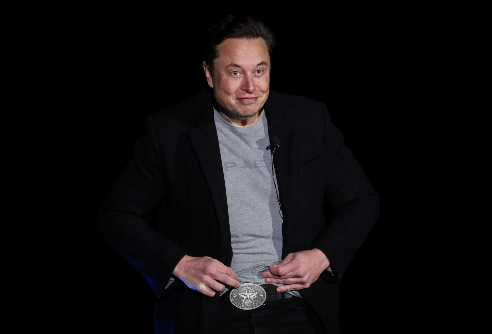 Musk, wearing a black blazer and a gray SpaceX T-shirt, lifts the hem of his shirt to show a large silver belt buckle featuring a five-pointed star with Tesla’s stylized “T” logo in the middle.