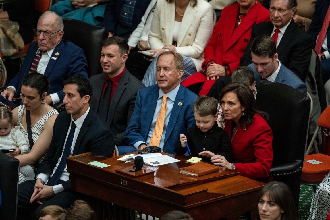 A man in a blue suit and a woman in a red suit sit together behind a wooden desk surrounded by people in folding chairs. The woman holds a child on her lap.