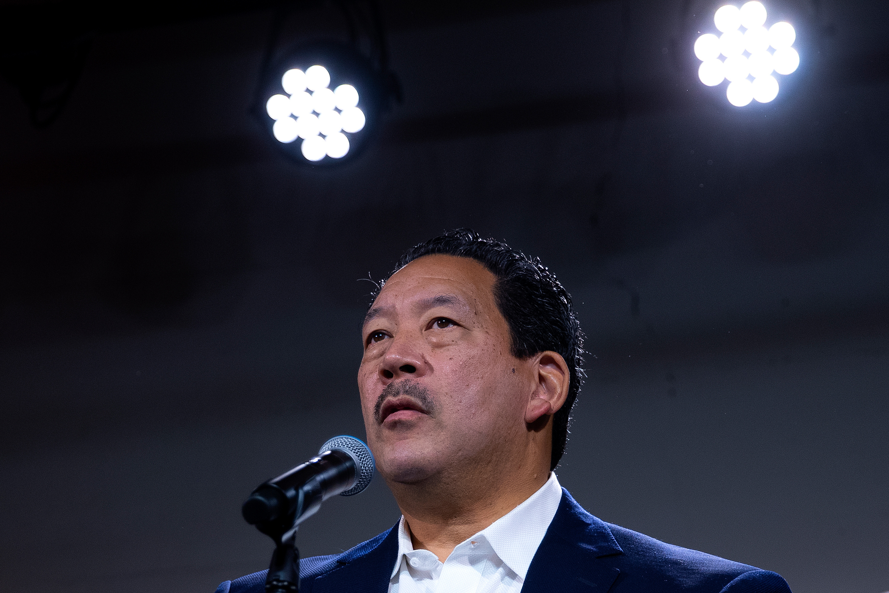 A man with black hair and a moustache, wearing a suit, stands at a microphone with two shining lights above him.