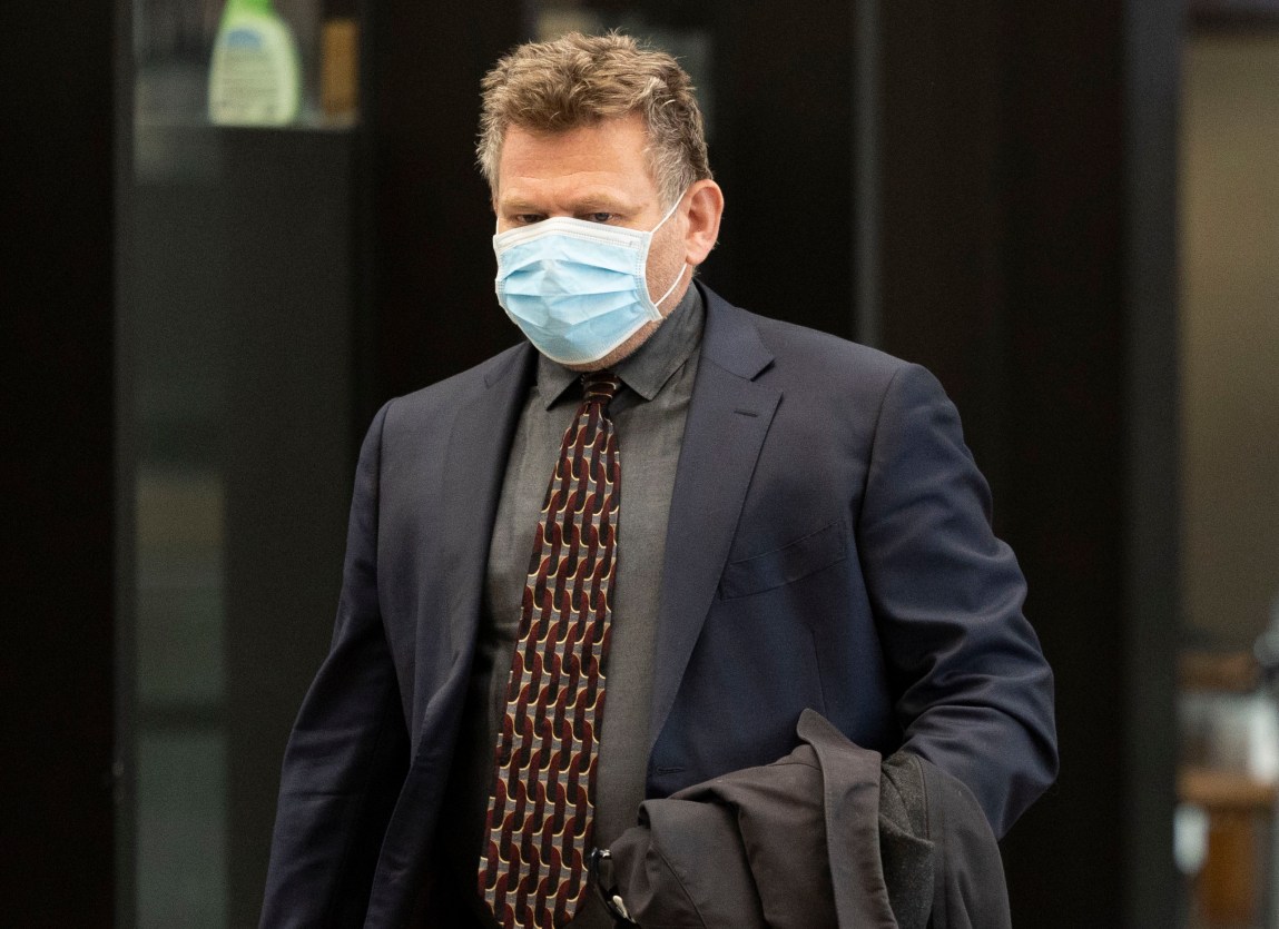 A man in a suit with a gray shirt. He has salt-and-pepper hair and is wearing a surgical mask.