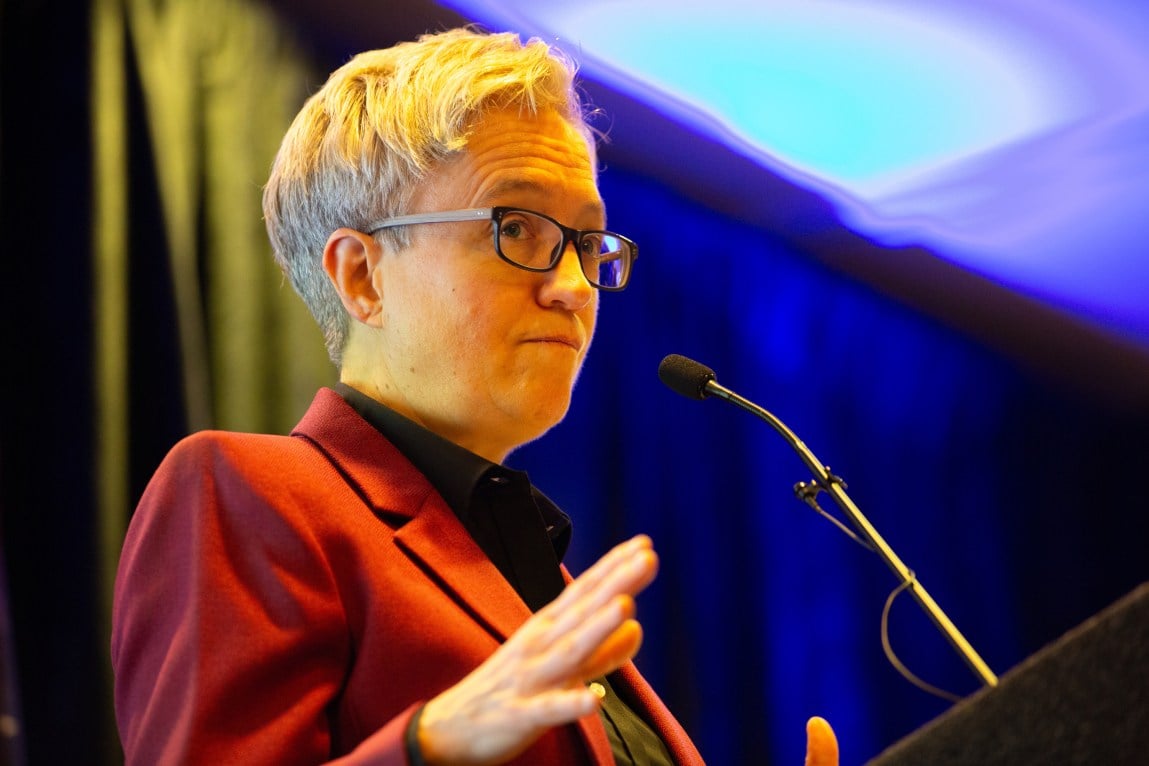 Tina Kotek, wearing glasses and a red blazer, stands at a podium.