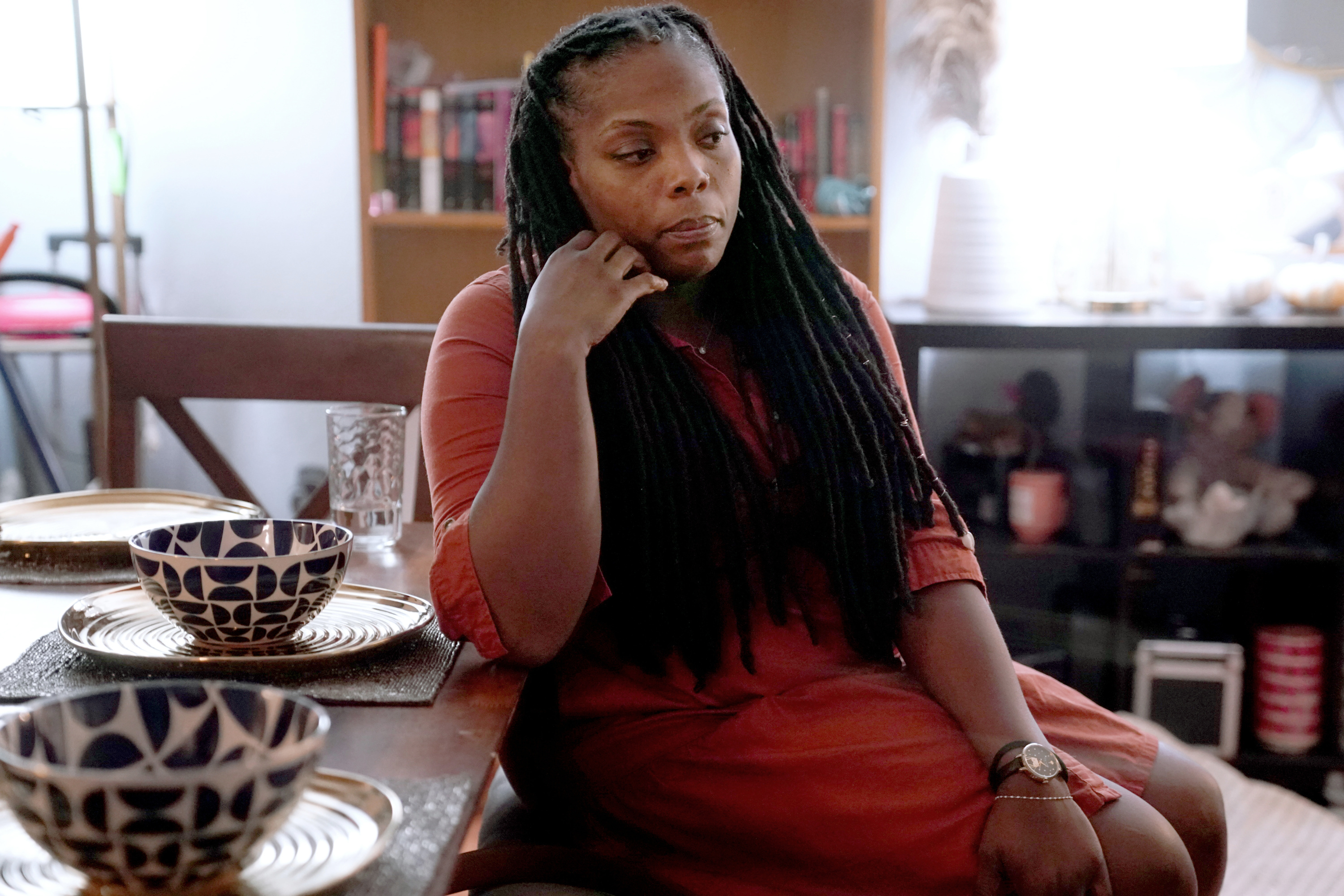 A woman in an orange dress sits in a dining room, with her head in her hand, staring off into the distance.