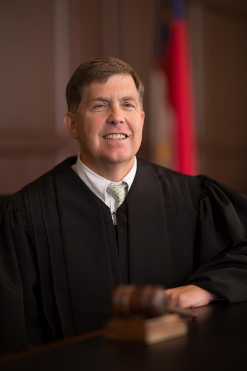 A man with brown hair smiles at the camera while wearing judges’ robes.