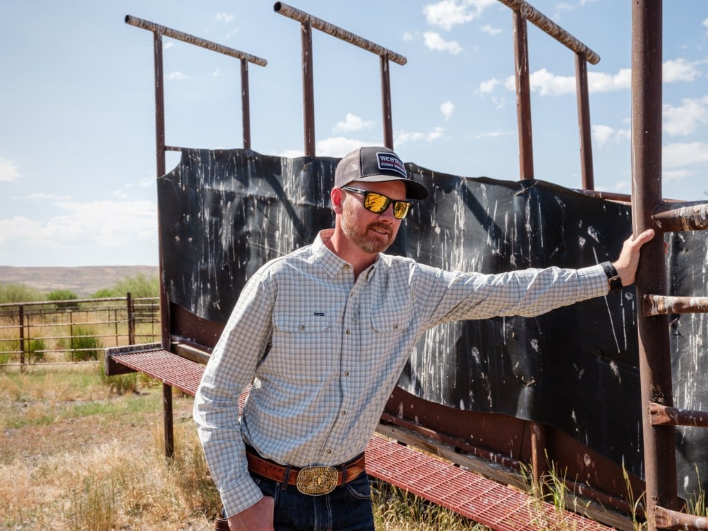A man wearing a baseball hat, sunglasses, a button-up shirt and jeans, with a prominent belt buckle, leans on a cattle chute.