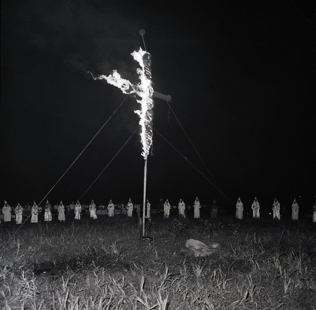 A circle of people in white robes surrounding a large burning cross that is propped up in a field.