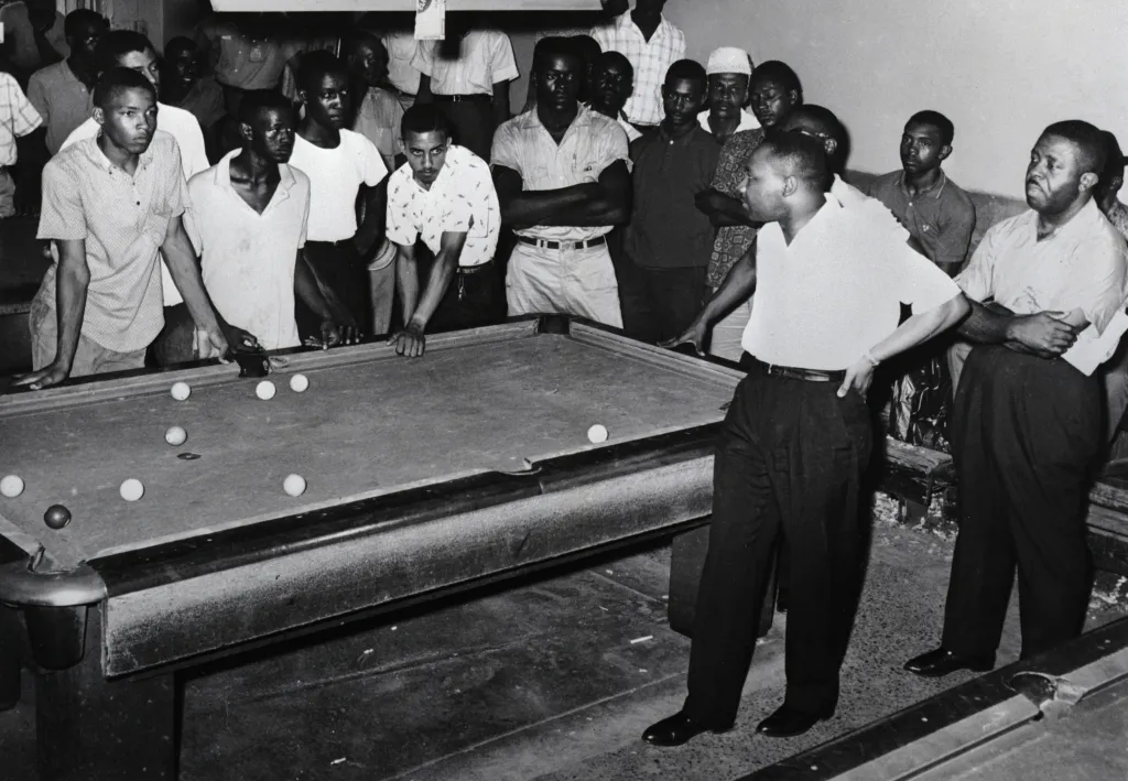 A group of men huddle around a pool table looking toward the Rev. Martin Luther King Jr. and the Rev. Ralph Abernathy.