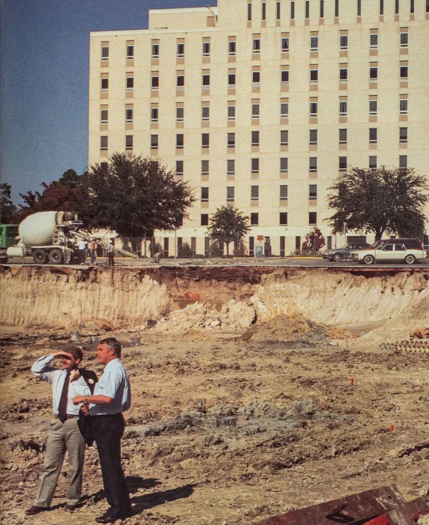 Two men stand together in a large dirt clearing. Behind them is a towering white building. The man on the left has his hand raised over his eyes blocking the sun.