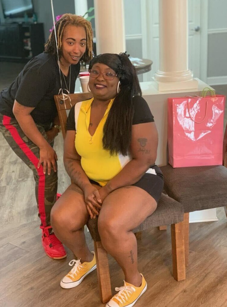 A woman is smiling and sitting in a chair while a second woman, also smiling, crouches behind her.
