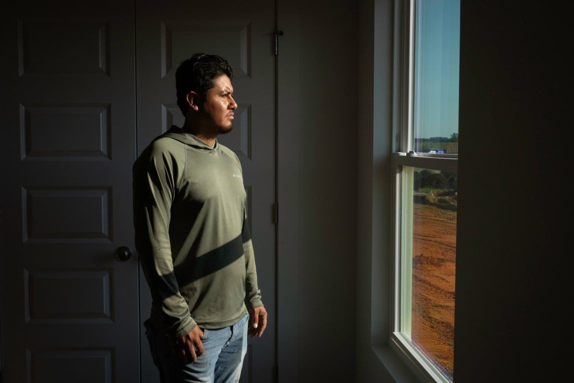 A man with a goatee and black hair wearing a green hoodie and jeans stands in a dark room looking out of a window that shows burnt sienna ground and blue sky.