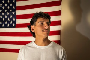 A man in a white T-shirt stands in front of an American flag.