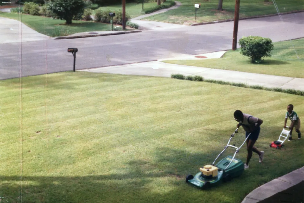 A man mowing a green front lawn, and a little boy is following him with a small lawn mower. They are perfectly mimicking each other. They are in a suburban setting.