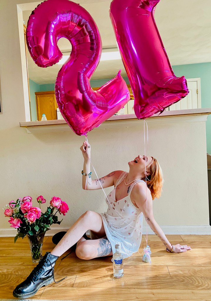 A young woman laughingly poses under pink balloons that spell out 21 in honor of her birthday.