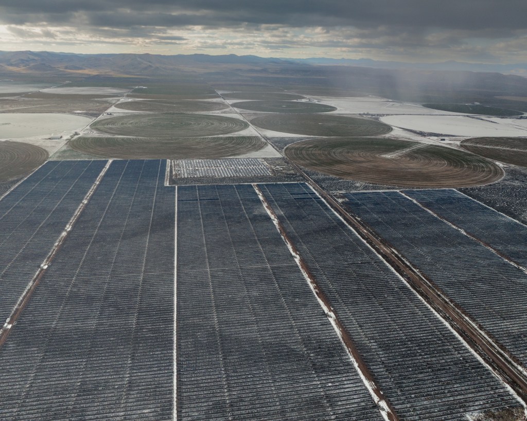 An aerial view of a large array of solar panels and center-pivot irrigation circles, lightly dusted with snow, under a cloudy sky, with mountains in the far distance.