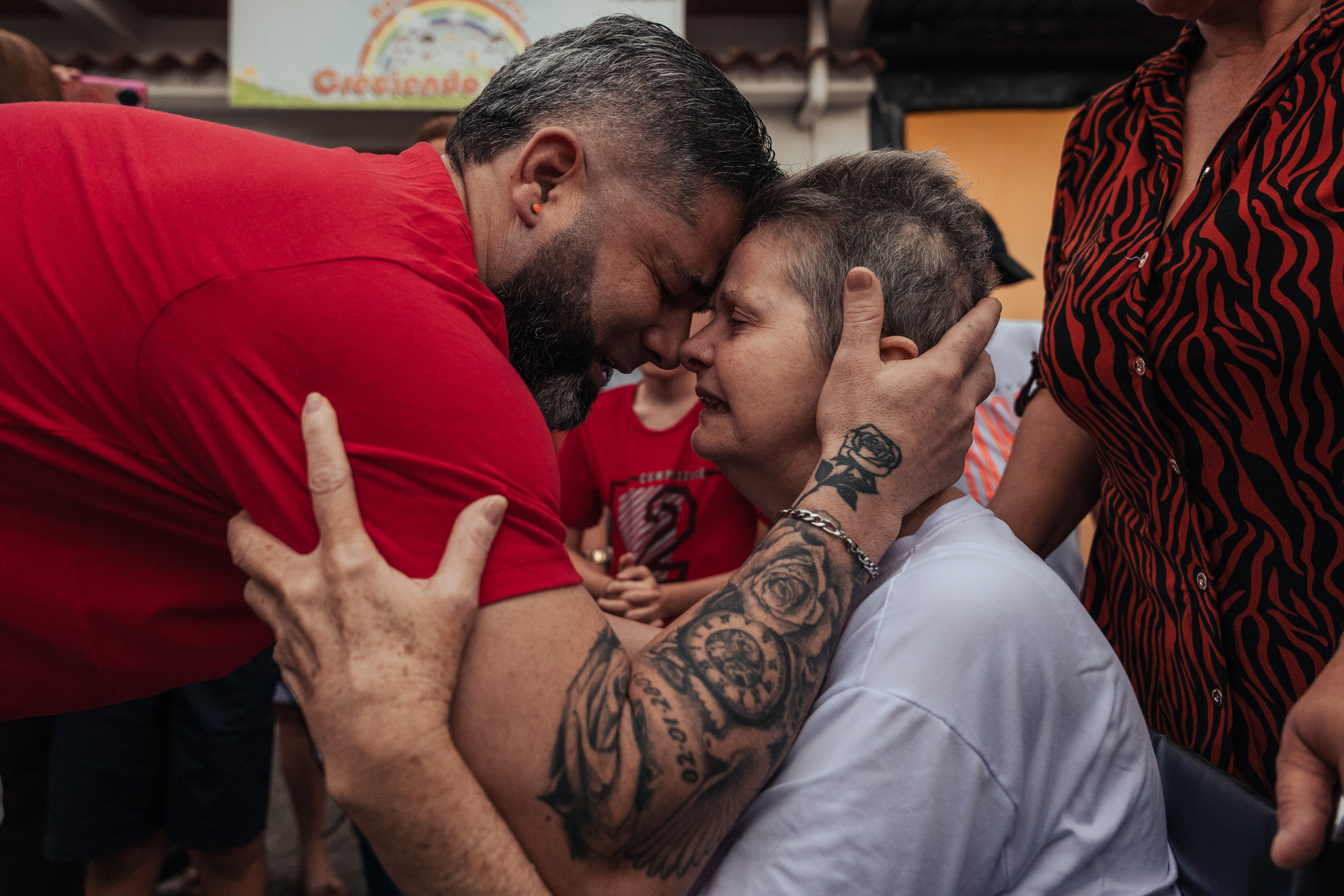 A man in jeans and a red T-shirt kneels in a road with his arm around a woman in jeans and a pink cardigan.