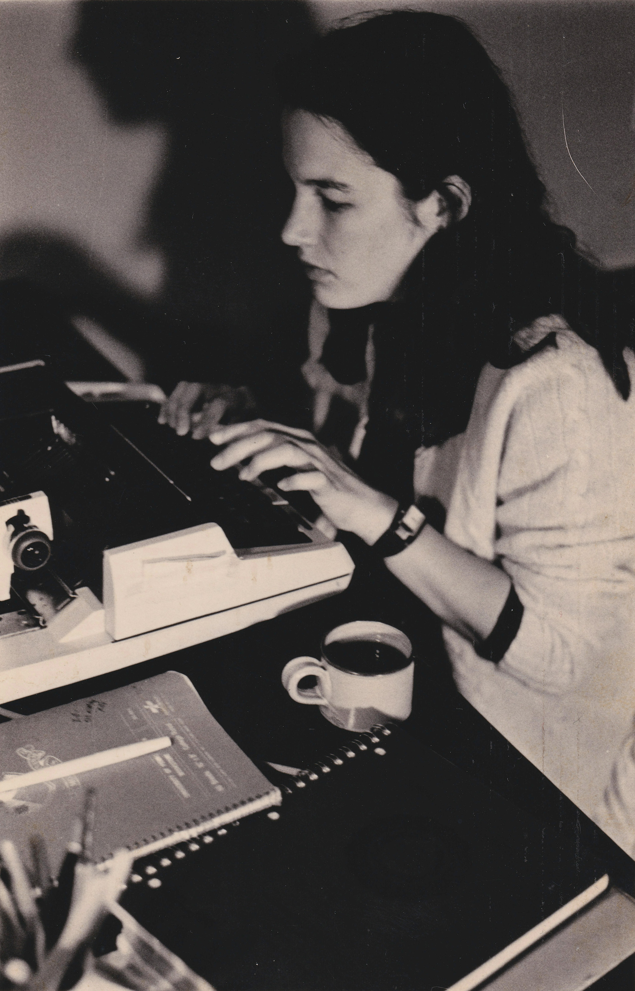 A young woman with black hair uses a typewriter at a desk. Beside her are a cup of coffee and notebooks.